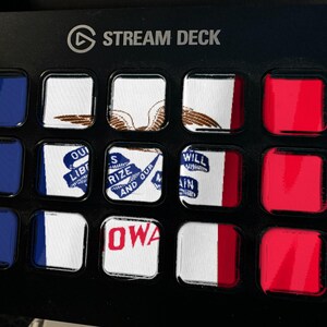 Puede incluir: Un dispositivo Stream Deck negro con quince botones cuadrados dispuestos en una cuadr&iacute;cula. Los botones muestran varios segmentos de banderas y texto, incluida la palabra IOWA, junto a botones de color azul y rojo s&oacute;lido a los lados.