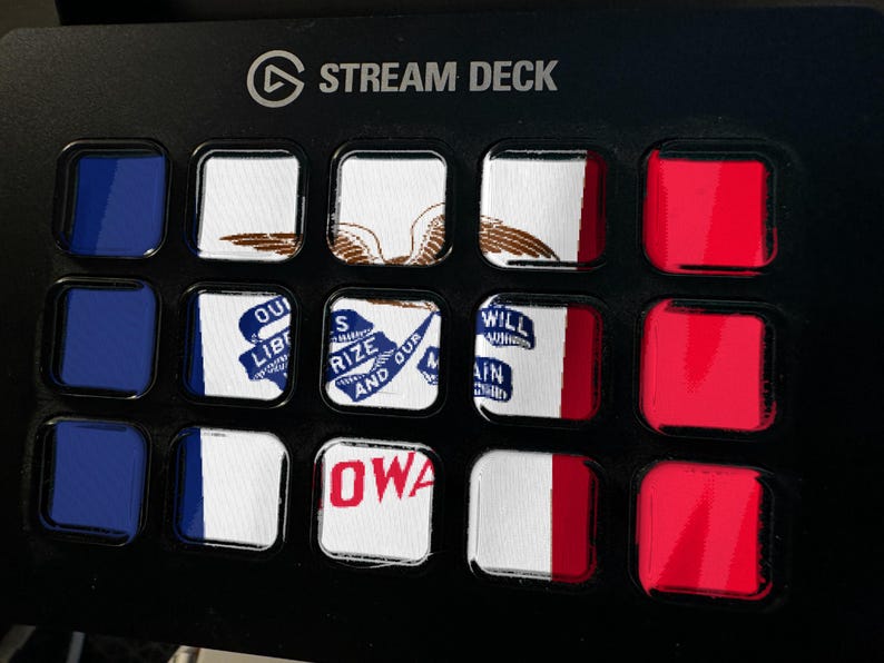 Puede incluir: Un dispositivo Stream Deck negro con quince botones cuadrados dispuestos en una cuadr&iacute;cula. Los botones muestran varios segmentos de banderas y texto, incluida la palabra IOWA, junto a botones de color azul y rojo s&oacute;lido a los lados.