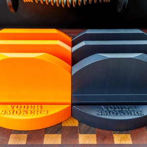 May include: A split image showcases two sets of grill accessories. On the left, four bright orange grill stands are visible, while the right side features four black stands. Each stand has the text "YODER SMOKERS" embossed on the base.