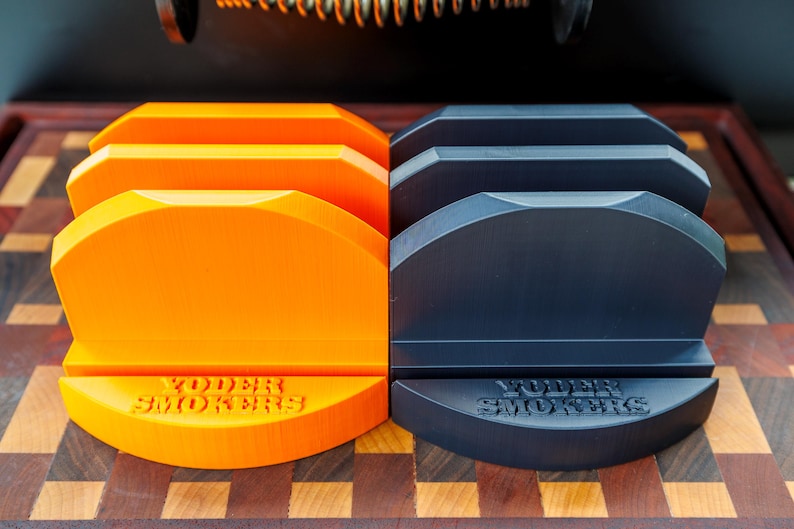 Op de afbeelding: De afbeelding toont twee sets barbecue-accessoires. Links staan vier oranje grillstandaards en rechts vier zwarte standaards. Elke standaard heeft de tekst "YODER SMOKERS" in reli&euml;f op de basis.