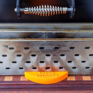 Op de afbeelding: Close-up van een smoker met een roestvrijstalen behuizing en een veergreep. De smoker heeft een geperforeerd ontwerp en een oranje, halfrond stuk aan de onderkant met de tekst "Yoder Smokers". De smoker staat op een houten oppervlak.