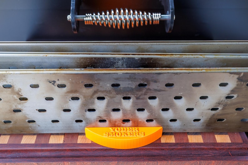 Op de afbeelding: Close-up van een smoker met een roestvrijstalen behuizing en een veergreep. De smoker heeft een geperforeerd ontwerp en een oranje, halfrond stuk aan de onderkant met de tekst "Yoder Smokers". De smoker staat op een houten oppervlak.