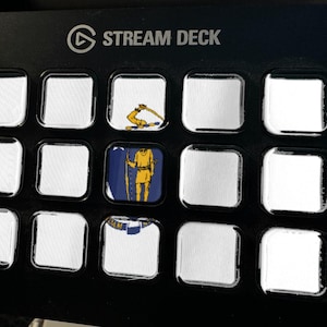 May include: A black Stream Deck control panel with fifteen square keys. Three keys feature printed graphics: one shows a yellow arm holding a sword, one displays a yellow figure on a blue background, and one shows a partial blue and white emblem.