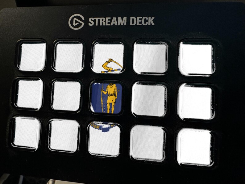 May include: A black Stream Deck control panel with fifteen square keys. Three keys feature printed graphics: one shows a yellow arm holding a sword, one displays a yellow figure on a blue background, and one shows a partial blue and white emblem.