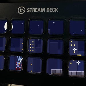 Może przedstawiać: Czarna konsola sterująca Stream Deck z piętnastoma kwadratowymi klawiszami LCD. Klawisze wyświetlają ciągłą scenę w stylu pixel art przedstawiającą nocny krajobraz miejski z księżycem, gwiazdami, oknami budynk&oacute;w i postacią z mieczem.