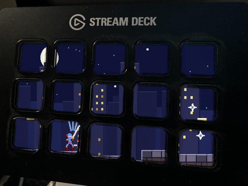 Może przedstawiać: Czarna konsola sterująca Stream Deck z piętnastoma kwadratowymi klawiszami LCD. Klawisze wyświetlają ciągłą scenę w stylu pixel art przedstawiającą nocny krajobraz miejski z księżycem, gwiazdami, oknami budynk&oacute;w i postacią z mieczem.