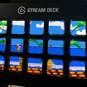 Puede incluir: Un dispositivo Stream Deck negro con quince teclas LCD cuadradas dispuestas en una cuadr&iacute;cula. Cada tecla muestra un segmento de una escena de playa tropical pixelada con agua azul, palmeras, nubes y un personaje, formando un paisaje.