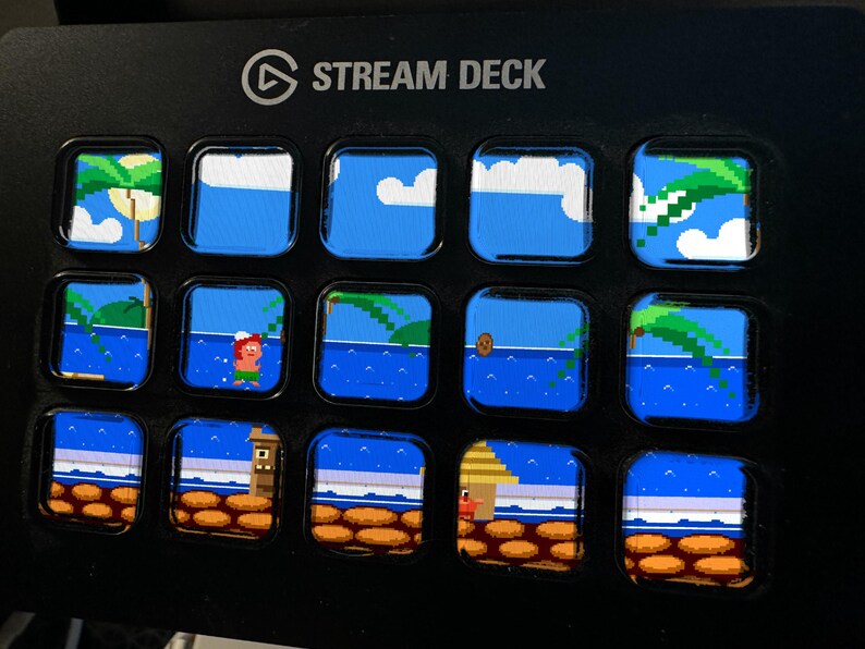 Puede incluir: Un dispositivo Stream Deck negro con quince teclas LCD cuadradas dispuestas en una cuadr&iacute;cula. Cada tecla muestra un segmento de una escena de playa tropical pixelada con agua azul, palmeras, nubes y un personaje, formando un paisaje.