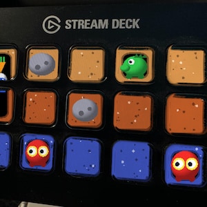 May include: A black Stream Deck control console featuring a grid of fifteen square buttons. The buttons display various pixelated icons, including a white creature with a carrot, grey spheres, a green creature, and red ghost-like figures on blue backgrounds.