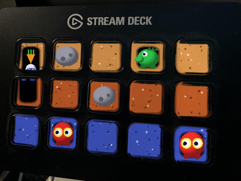 May include: A black Stream Deck control console featuring a grid of fifteen square buttons. The buttons display various pixelated icons, including a white creature with a carrot, grey spheres, a green creature, and red ghost-like figures on blue backgrounds.
