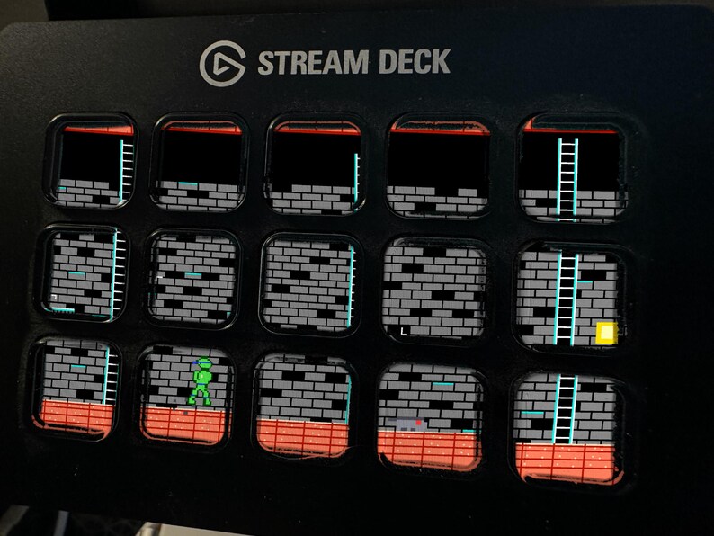 Op de afbeelding: Een zwart Stream Deck-apparaat met vijftien vierkante LCD-toetsen. Elke toets toont een segment van een retro 8-bit videospel-level, met grijze bakstenen muren, oranje vloertegels, blauwe ladders en een klein groen figuurtje.