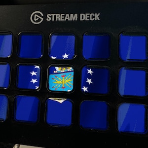 May include: A black Stream Deck control panel featuring a grid of fifteen square buttons. Most buttons display a solid dark blue colour, while three buttons show a blue flag with white stars and a central yellow emblem with a blue background.