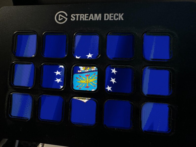 May include: A black Stream Deck control panel featuring a grid of fifteen square buttons. Most buttons display a solid dark blue colour, while three buttons show a blue flag with white stars and a central yellow emblem with a blue background.