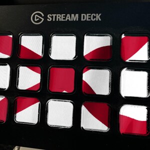 K&ouml;nnte beinhalten: Ein schwarzes Stream Deck Bedienfeld mit f&uuml;nfzehn quadratischen Tasten in einem Raster. Jede Taste zeigt ein rot-wei&szlig;es Grafikmuster. Der Text STREAM DECK ist in Wei&szlig; oben mittig auf dem schwarzen Geh&auml;use des Ger&auml;ts aufgedruckt.