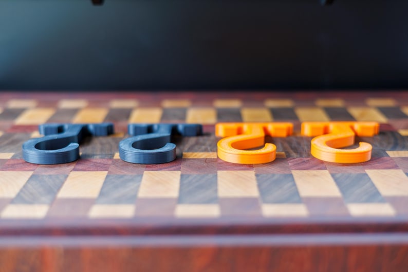 May include: Four 3D-printed hooks in a row. Two are dark gray, and two are bright orange. The hooks are on a wooden surface with a checkerboard pattern of various wood tones.