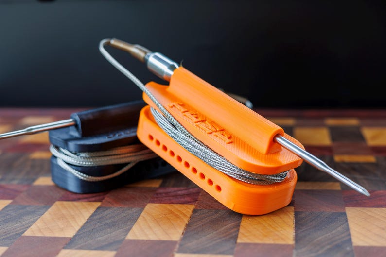 May include: Two temperature probe holders, one black and one bright orange, with stainless steel probes and coiled wires. The orange holder has the word "FASA" printed on it. The holders are designed to keep the probes organized.
