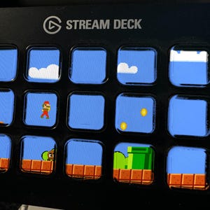 May include: A black Stream Deck controller featuring fifteen square LCD keys. Each key displays a pixelated video game scene with blue skies, white clouds, brown brick platforms, gold coins, a green pipe, and a character in red and brown clothing.