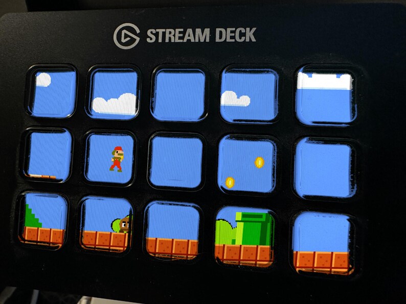 May include: A black Stream Deck controller featuring fifteen square LCD keys. Each key displays a pixelated video game scene with blue skies, white clouds, brown brick platforms, gold coins, a green pipe, and a character in red and brown clothing.