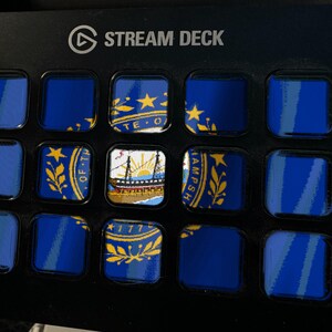 May include: A black Stream Deck control console featuring a grid of fifteen square LCD keys. The keys display a fragmented image of the New Hampshire state flag, including the state seal, a ship, and the year 1776, set against a dark background.