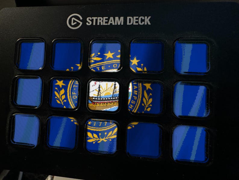 May include: A black Stream Deck control console featuring a grid of fifteen square LCD keys. The keys display a fragmented image of the New Hampshire state flag, including the state seal, a ship, and the year 1776, set against a dark background.