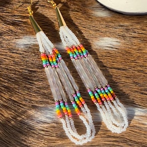 May include: A pair of long, beaded earrings with gold-colored hooks. The earrings feature clear, elongated beads with sections of colorful rainbow beads. The bottom of the earrings have multiple strands of white beads.