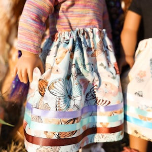 May include: A doll wearing a colorful striped long-sleeved shirt and a light blue skirt adorned with butterfly patterns. The skirt is trimmed with ribbons in shades of purple, teal, and brown. The doll is wearing white sneakers.