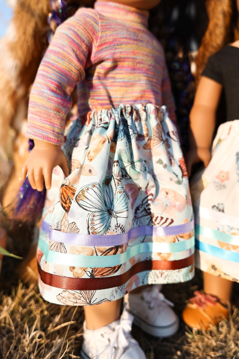 May include: A doll wearing a colorful striped long-sleeved shirt and a light blue skirt adorned with butterfly patterns. The skirt is trimmed with ribbons in shades of purple, teal, and brown. The doll is wearing white sneakers.