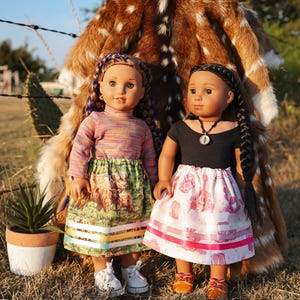 May include: Two dolls dressed in colorful outfits stand in front of a deer hide. One doll wears a striped top and a skirt with a Highland cow print. The other doll is in a black top and a pink skirt with a pattern. Both dolls have braided hair.
