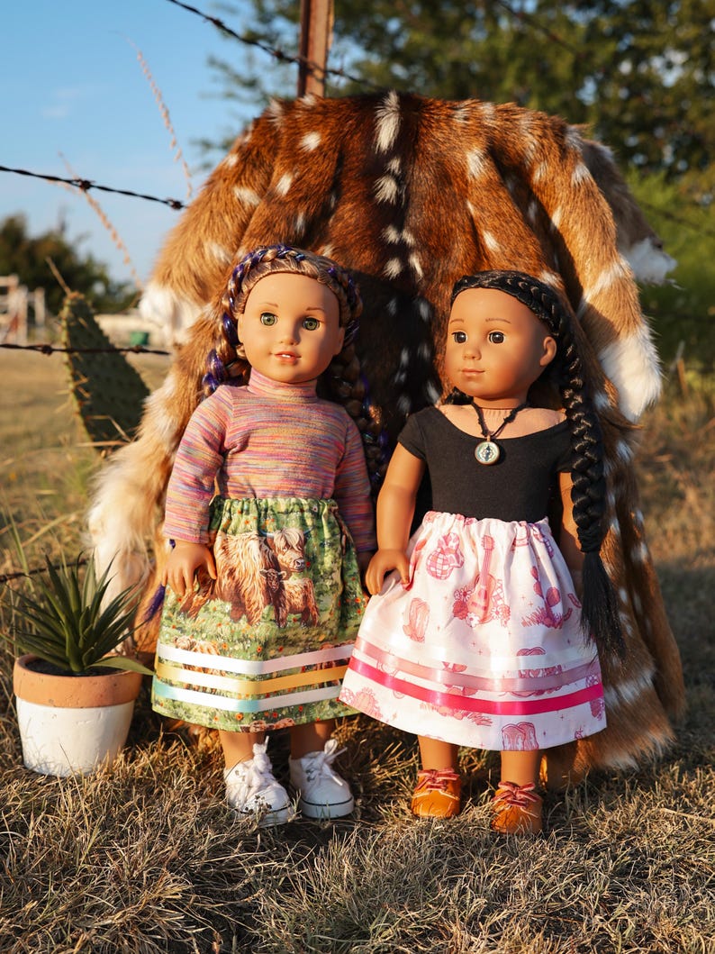 May include: Two dolls dressed in colorful outfits stand in front of a deer hide. One doll wears a striped top and a skirt with a Highland cow print. The other doll is in a black top and a pink skirt with a pattern. Both dolls have braided hair.