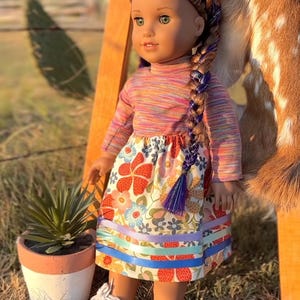 May include: A doll with green eyes and a long, braided hairstyle. The doll wears a long-sleeved, striped shirt and a floral skirt with ribbon trim. White sneakers complete the outfit. A small potted plant sits nearby.