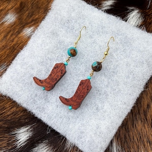 May include: A pair of gold-colored dangle earrings. Each earring features a small, brown cowboy boot charm with a decorative design, a turquoise bead, and a brown and turquoise bead. The earrings are displayed on a white surface with a brown and white background.