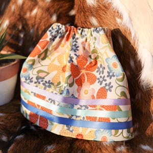 May include: A floral print fabric item with orange, yellow, and blue flowers, and green leaves. The item has a gathered top and is adorned with horizontal ribbons in lavender, light blue, and dark blue. The item is set against a brown and white spotted background.