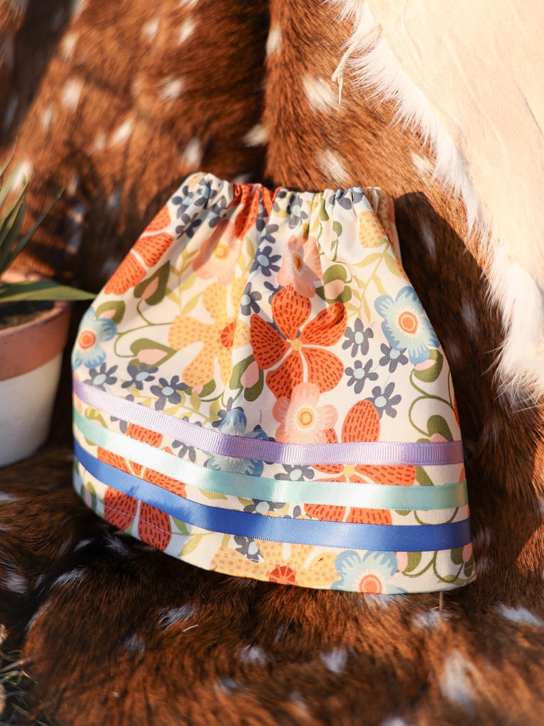 May include: A floral print fabric item with orange, yellow, and blue flowers, and green leaves. The item has a gathered top and is adorned with horizontal ribbons in lavender, light blue, and dark blue. The item is set against a brown and white spotted background.