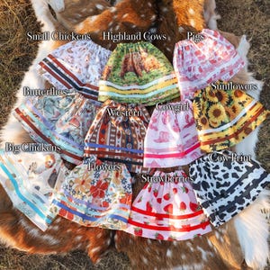 May include: A collection of colorful, patterned skirts with various designs, including chickens, highland cows, pigs, butterflies, sunflowers, cowgirl, western, flowers, strawberries, and cow print. Each skirt has a contrasting band at the hem and waist.
