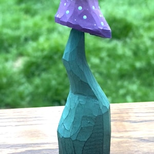 May include: A carved wooden mushroom sculpture. The mushroom cap is purple with light blue dots, and the stem is teal. The sculpture is on a wooden surface with a green background.