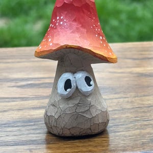 May include: A whimsical wooden mushroom figurine with a red cap dotted with white spots and an orange rim. The mushroom has a light brown stem with large, expressive eyes. The carving has a textured, rustic appearance.