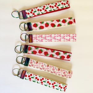 May include: A collection of six keychains with various patterns. The keychains feature designs of cherries, strawberries, bows, and candy. Each keychain has a metal ring and a colorful, iridescent clasp.