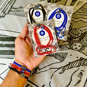 May include: Three evil eye bracelets with silver charms. The bracelets are red, blue, and black string. The bracelets are on a white background with a floral pattern.