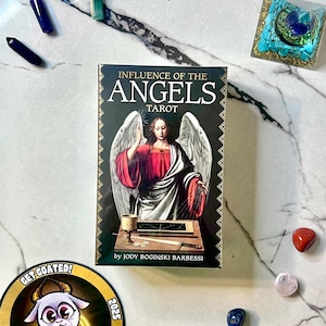 May include: A tarot deck titled "Influence of the Angels" with an illustration of an angel on the cover. The image includes crystals, a sticker with a goat graphic, and other decorative elements on a marble surface.