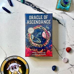 May include: A deck of "Oracle of Ascendance" cards with a cosmic design, featuring a snake encircling planets. The image includes crystals, a decorative pyramid, and a "Get Goated!" sticker with a goat illustration. The background is a white marble surface.