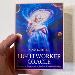 May include: A rectangular box featuring the "LIGHTWORKER ORACLE" by Alana Fairchild. The cover art depicts a woman in a flowing gown within a swirling galaxy, with planets in the background. The text reads "Guidance & Empowerment for those Who Love the Light."