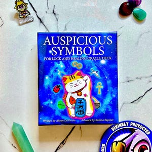 May include: A square oracle deck box titled Auspicious Symbols for Luck and Healing Oracle Deck. The blue cover features a white and orange beckoning cat illustration, a ladybug, an ankh, and coins. Written by Alison DeNicola, artwork by Sabina Espinet.
