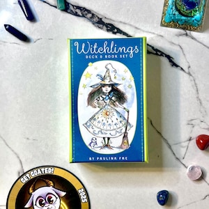 May include: A "Witchlings Deck & Book Set" box with a blue and yellow design, featuring a witch illustration. The image includes crystals, a sticker with a goat, and other decorative items on a marble surface.