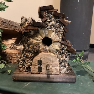 May include: A birdhouse crafted from wood and stone, featuring a small door and a circular opening. The birdhouse is adorned with pebbles and twigs.