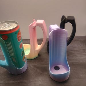 May include: A collection of 3D-printed can holders in various colors. One holder is holding a can of soda. The holders have a handle and a base to secure the can. Colors include blue, pink, purple, and black.
