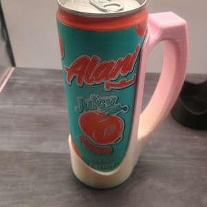 May include: A teal and orange can of 'Alan' Juicy Peach Energy drink, held in a pink 3D-printed mug-style holder. The can features a peach graphic and the word 'Energy'. The holder has a handle for easy gripping.