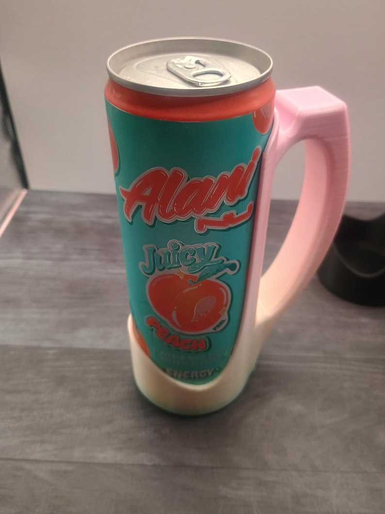 May include: A teal and orange can of 'Alan' Juicy Peach Energy drink, held in a pink 3D-printed mug-style holder. The can features a peach graphic and the word 'Energy'. The holder has a handle for easy gripping.