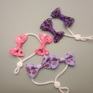 May include: Three hair accessories featuring small fabric bows attached to white elastic loops with adjustable white toggles. One bow has a purple leopard print, one is pink with white polka dots, and one is light purple with a pink daisy floral pattern.