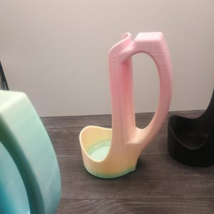 May include: A 3D-printed ladle rest in a gradient of pink, yellow, and green. The ladle rest has a curved handle and a small bowl-shaped base. Other similar items are visible in the background.