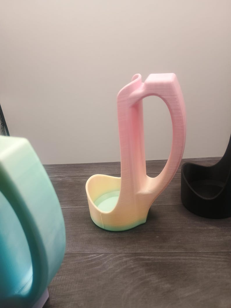 May include: A 3D-printed ladle rest in a gradient of pink, yellow, and green. The ladle rest has a curved handle and a small bowl-shaped base. Other similar items are visible in the background.